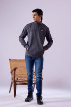 Person wearing a gray jacket, blue jeans, and black boots standing next to a wicker chair on a white background