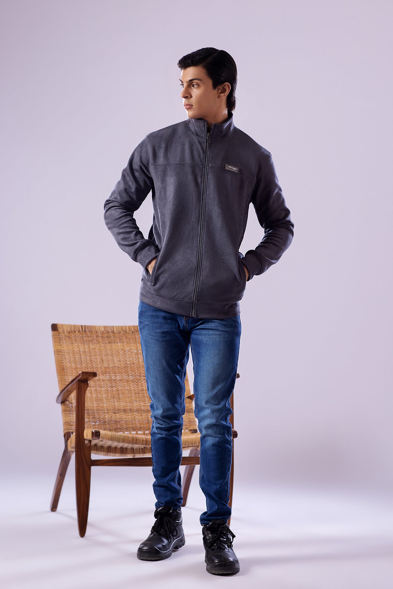 Person wearing a gray jacket, blue jeans, and black boots standing next to a wicker chair on a white background