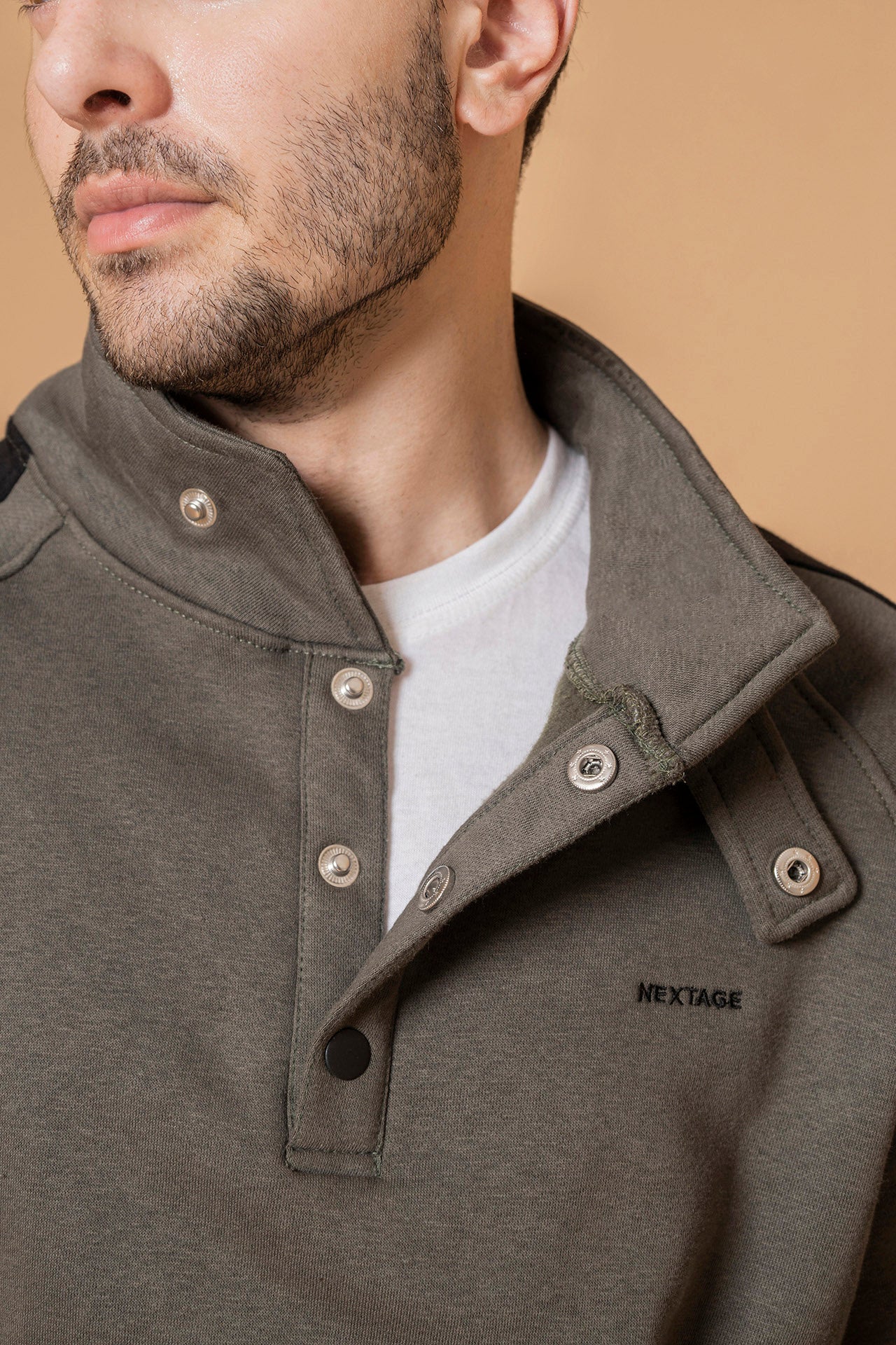 Close-up of nextage model wearing a green olive jacket with 'Nextage' branding on a beige background
