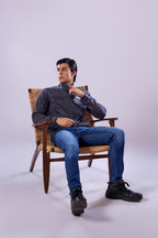 Man sitting on a wooden chair wearing a dark shirt, blue jeans, and black shoes against a white background