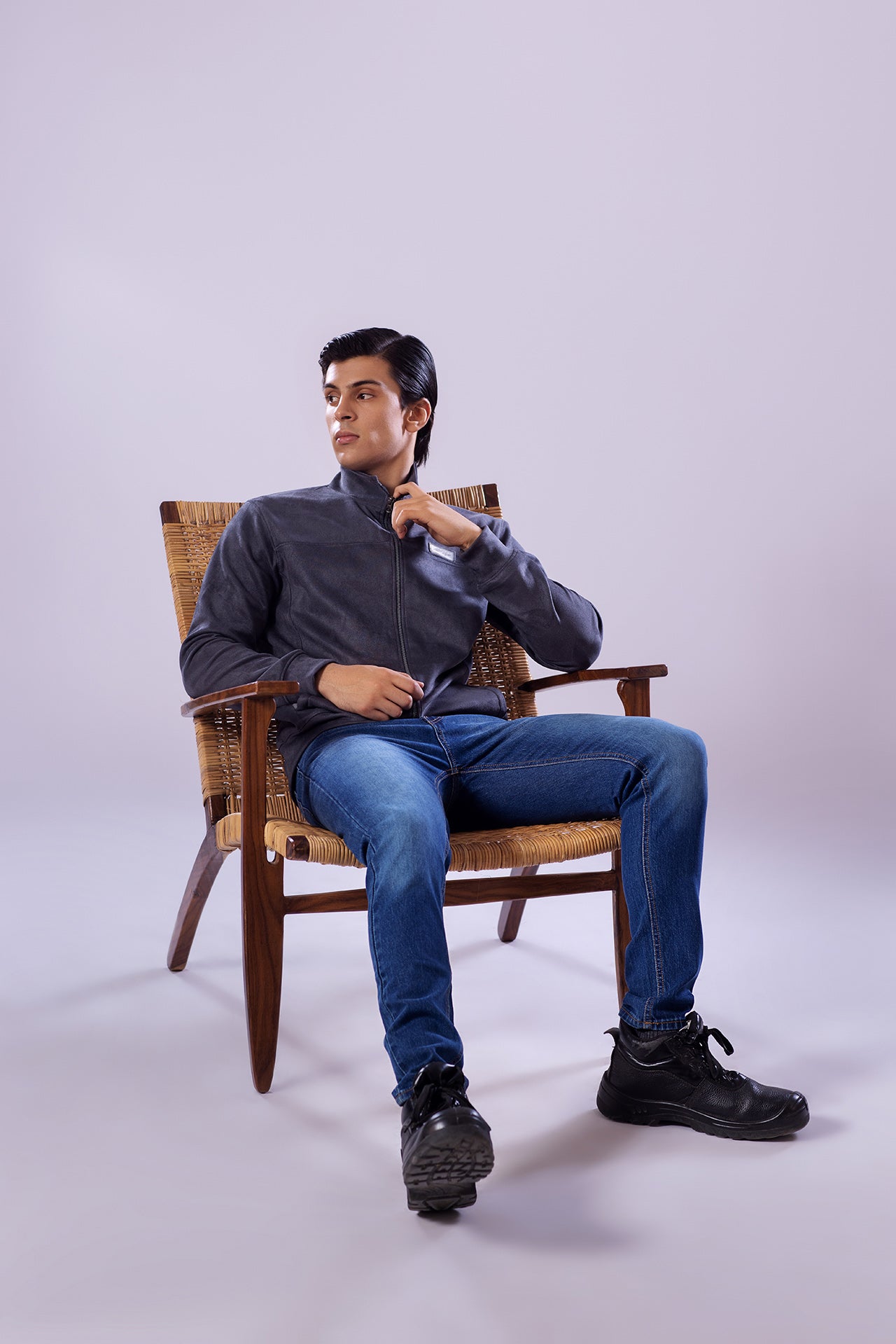 Man sitting on a wooden chair wearing a dark shirt, blue jeans, and black shoes against a white background