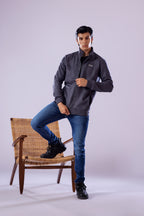 Man wearing a gray jacket and blue jeans sitting on a wooden chair against a white background