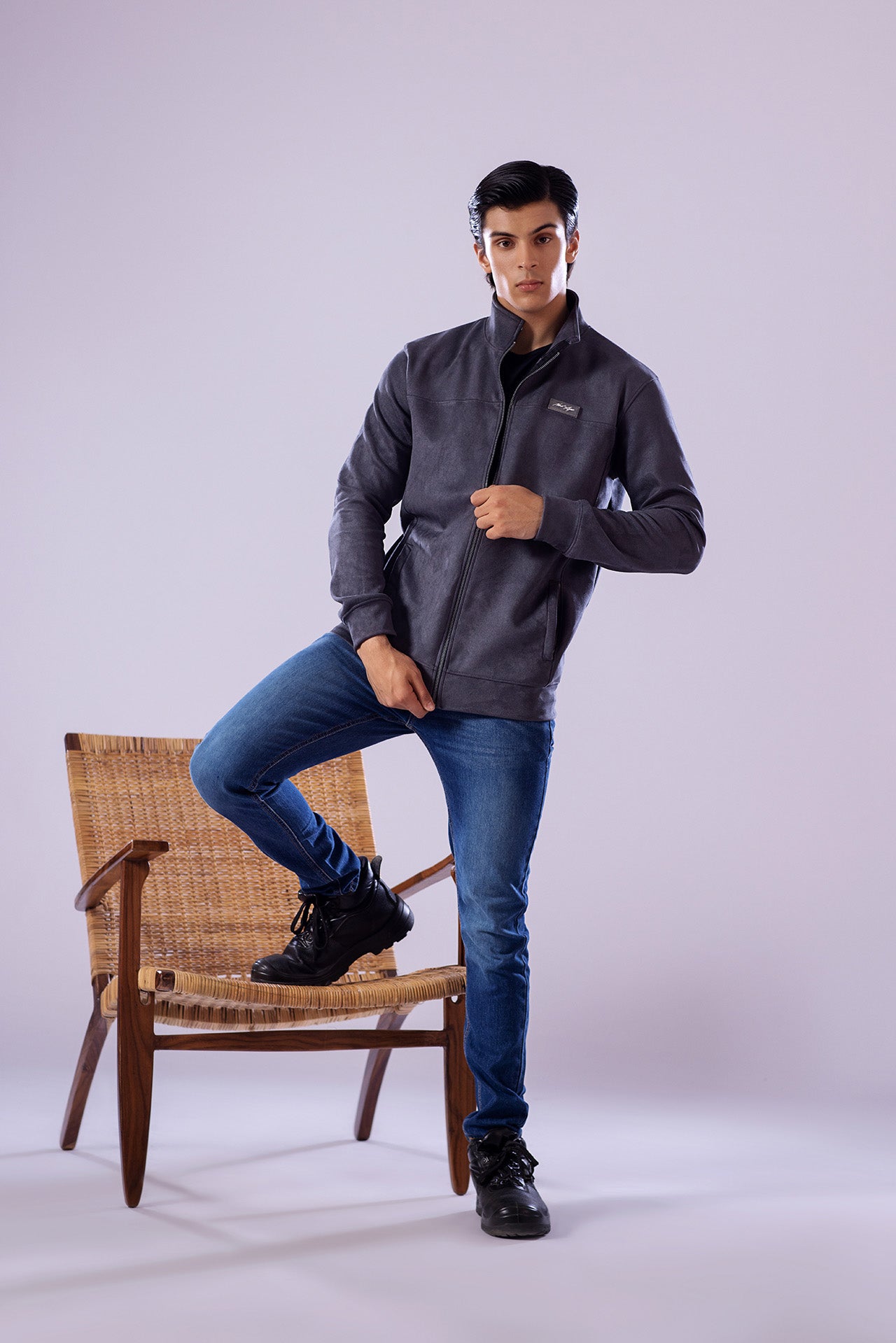 Man wearing a gray jacket and blue jeans sitting on a wooden chair against a white background